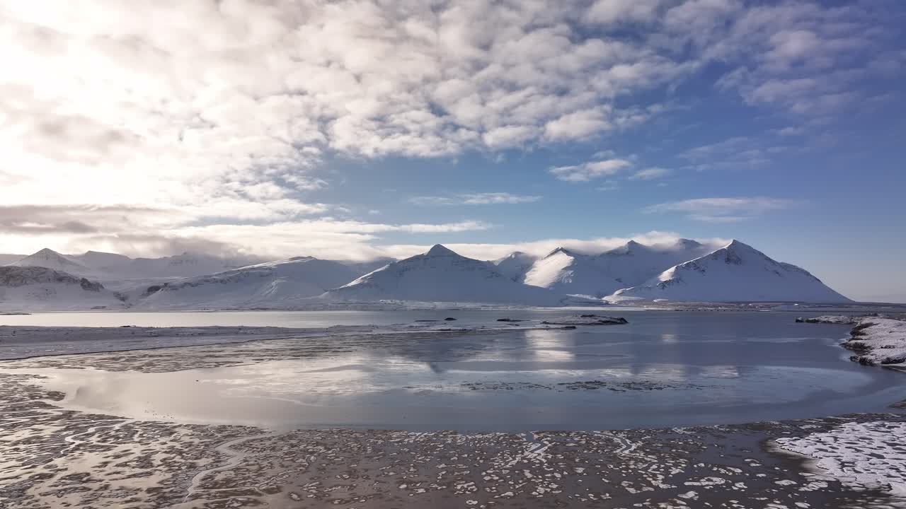 Winter aerial view of Borgarfjörður with Borgarnes town and Hafnarfjall mountain range. Snow-covered peaks reflect in the calm icy waters under a dramatic sky in west Iceland.