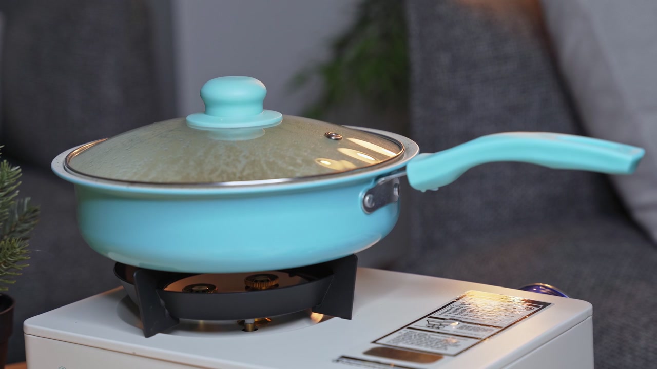 Close-up of a light blue frying pan with a steaming glass lid cooking on a white portable gas stove. The indoor domestic scene features a shallow depth of field with a gray sofa background.