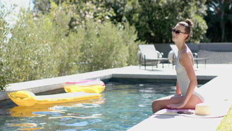 a teenage caucasian girl with brown hair tied back is sitting poolside with copy space