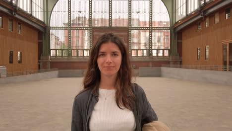 front-facing portrait of a young woman standing in a spacious indoor setting. she looks directly at the camera with a calm and confident expression, wearing casual clothes in neutral tones.