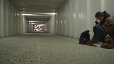 a girl in a brown coat sits bent over in an underpass tunnel, holding her phone with a saddened expression. the empty tunnel and her posture convey a deep sense of loneliness and emotional distress