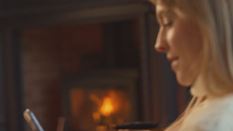 close up of woman at home in lounge with cosy fire and hot drink using mobile phone