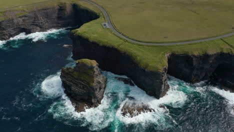 Luftaufnahme-Von-Kochendem-Wasser-An-Küstenfelsen.-Erstaunliche-Aufnahme-Von-Hohen-Klippen-Und-Grünem-Gras-Mit-Straße-An-Der-Spitze.-Kilkee-Cliff-Walk,-Irland