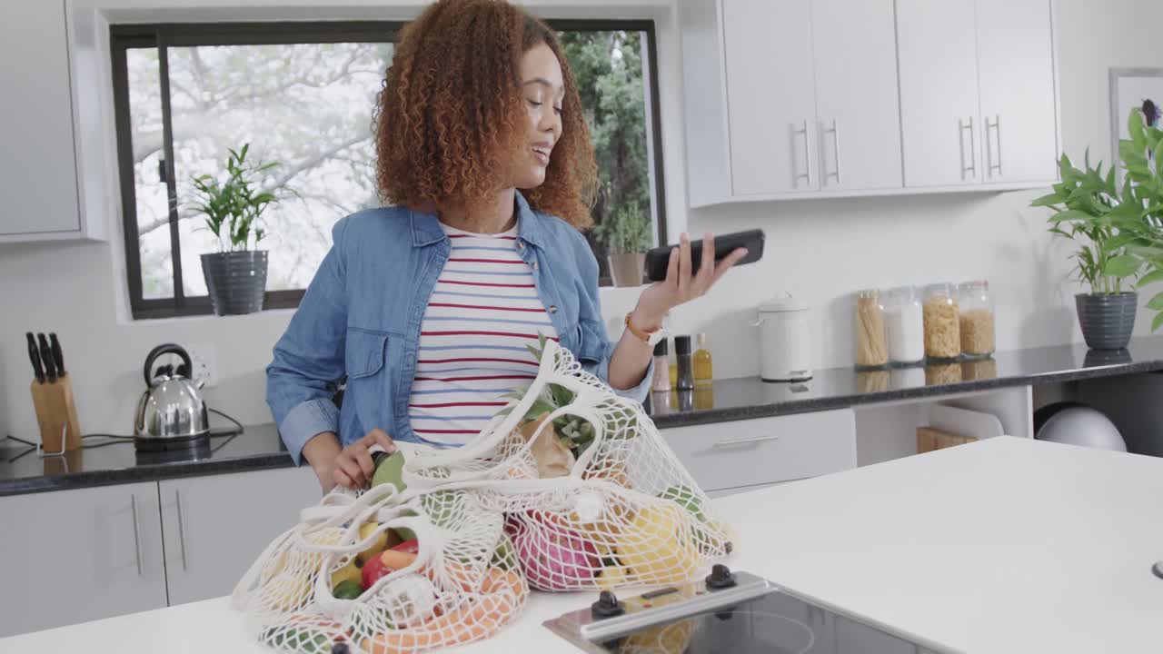 feliz mujer biracial hablando por teléfono y desempaquetando bolsas de comestibles en la cocina, cámara lenta