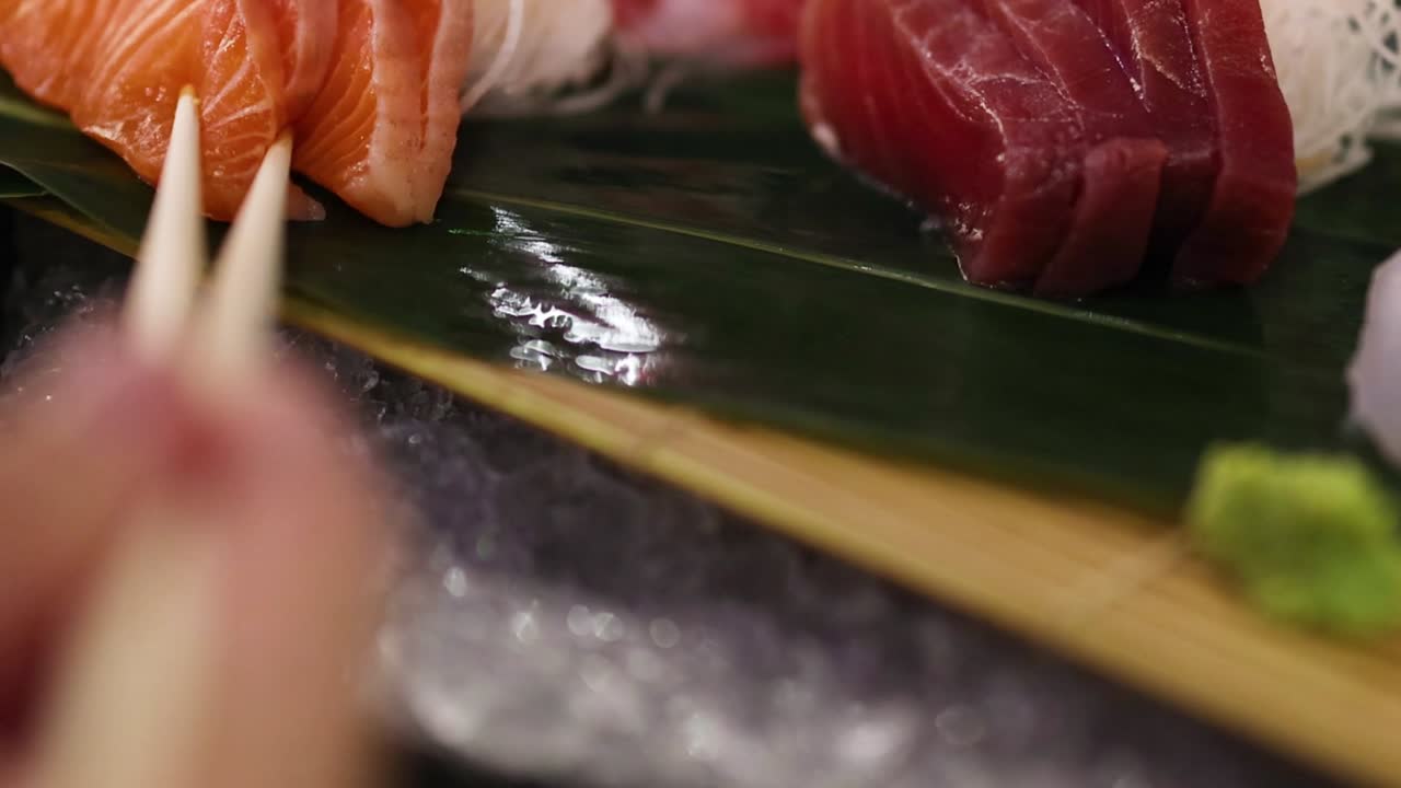 Primo piano di bacchette che prendono salmone e tonno da una foglia di bambù con guarnizione di wasabi.