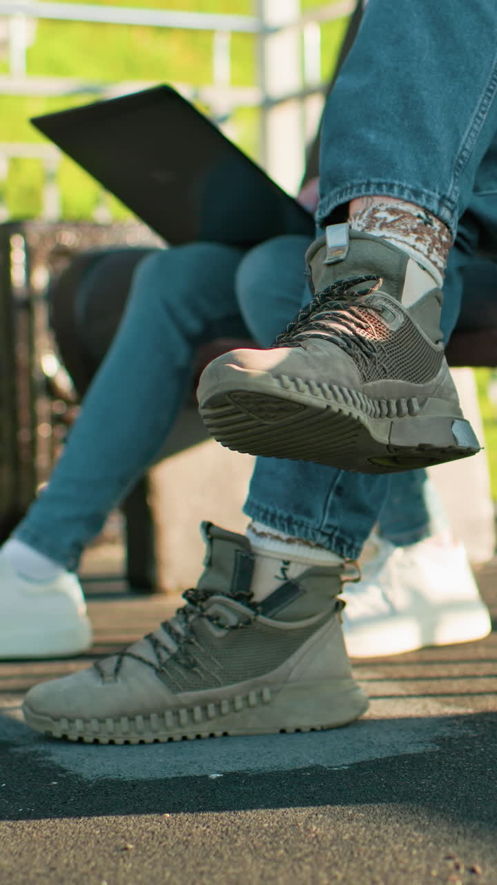 primer plano de las piernas de dos personas sentadas al aire libre en bancos de madera, con zapatillas deportivas y vaqueros casuales, una sosteniendo una computadora portátil mientras la otra muestra una mano tatuada, con sombras proyectadas por la luz del sol