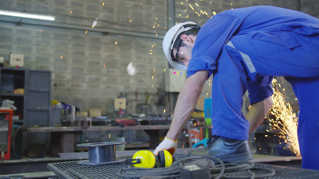 trabajador masculino caucásico en un uniforme seguro usar soldadores cueros, qc comprobación y medición con rueda eléctrica de molienda en la estructura de acero en el factor en el garaje, naranja bokeh chispas vuelan a los lados
