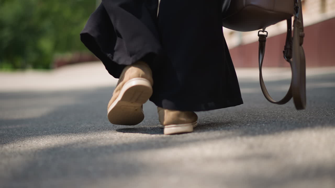 Back leg view of girl walking on sunny street carrying brown handbag with strap brushing pavement, sunlight and shadow adding depth to calm motion