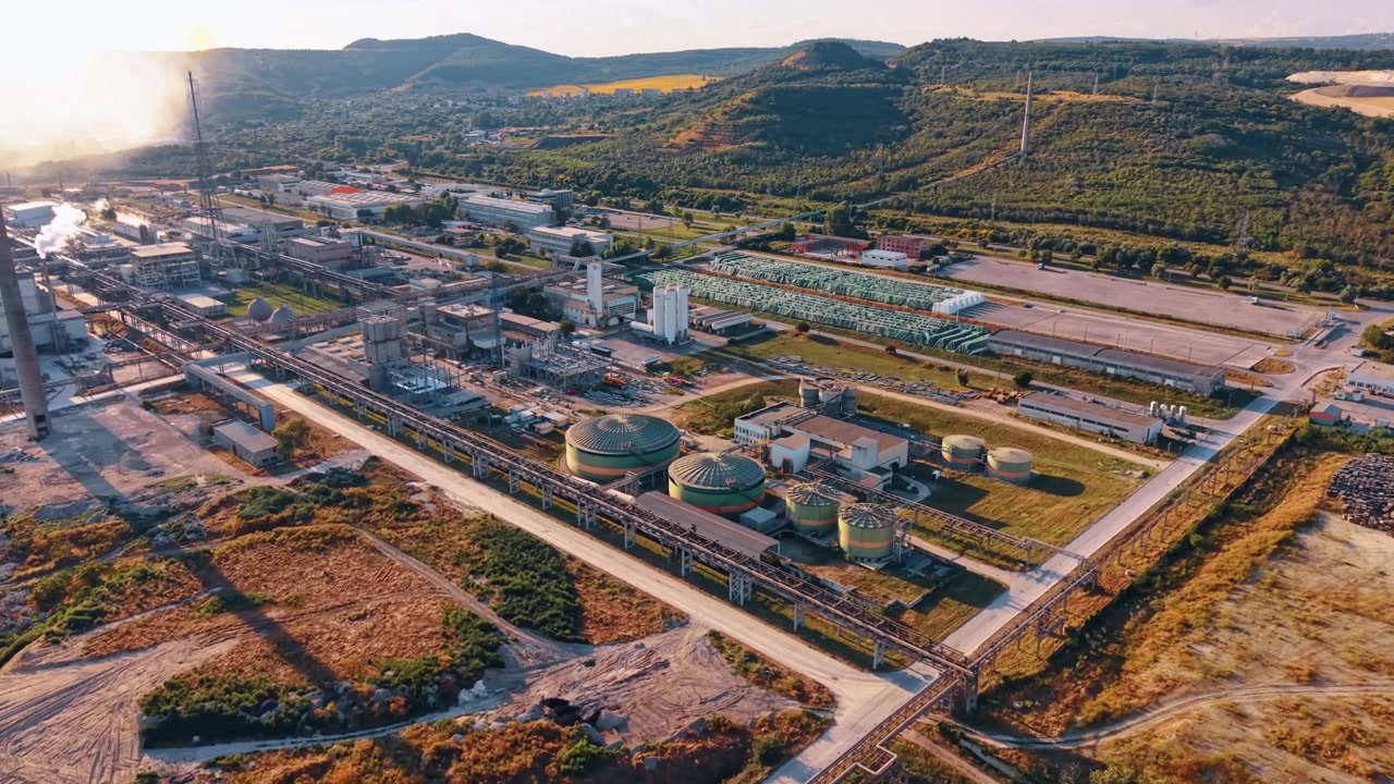 Chemical processing complex with tanks and elevated pipelines. Drone view of circular vessels, factory structures and pipe corridors with hills and quarry in background.
