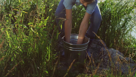 retrato de un lindo adolescente parado en la orilla del lago, sacando un pez de un balde después de pescar y mostrándolo a la cámara