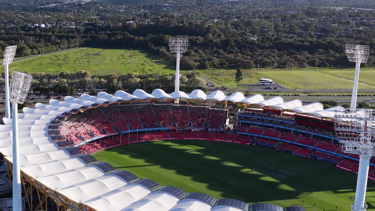 Drone pans across a large sports arena with red seating under bright daylight.
