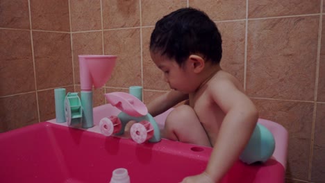 Un-Niño-Chino-Asiático-De-Tres-Años-Bañándose-Dentro-De-Una-Pequeña-Piscina-De-Plástico,-Llenando-Y-Vertiendo-Agua-De-Los-Contenedores,-Salpicando-Agua,-Observando-El-Agua-Con-La-Que-Está-Jugando