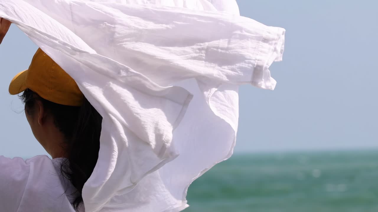 A person wearing a yellow cap unfurls a white fabric against a backdrop of the ocean.