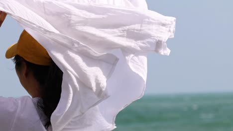 a person wearing a yellow cap unfurls a white fabric against a backdrop of the ocean.