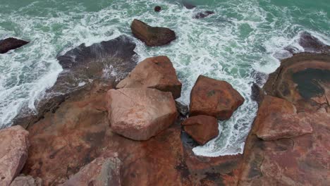 Stunning-aerial-forward-shot-of-a-reddish-rocky-coast-and-then-revealing-the-violent-and-intense-green-sea-with-splashing-waves