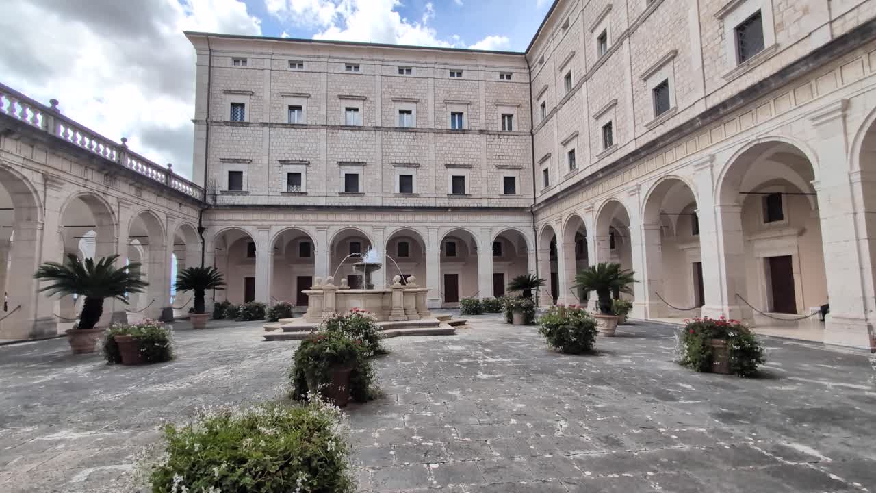 Slow pan across the courtyard of Monte Cassino Abbey in Italy, showing stone arches, historic walls, and a central fountain under soft daylight