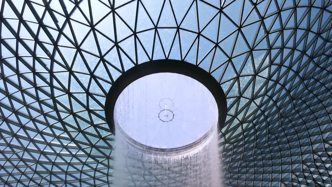 A vertical camera tilt reveals a large indoor waterfall surrounded by lush greenery beneath a geometric glass dome, with natural daylight illuminating the scene