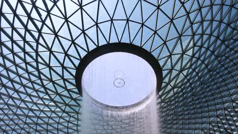 a vertical camera tilt reveals a large indoor waterfall surrounded by lush greenery beneath a geometric glass dome, with natural daylight illuminating the scene
