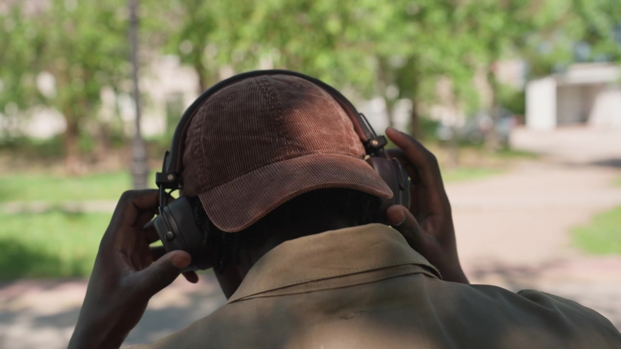 Close up of man wearing headphones and adjusting them in outdoor park setting with green trees pathway and soft daylight. subject is seen from behind as man interacts with headphone device
