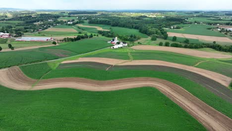 Luftaufnahme-Geschwungener-Landwirtschaftlicher-Felder-Mit-Wirtschaftsgebäuden,-Umgeben-Von-Viel-Grün