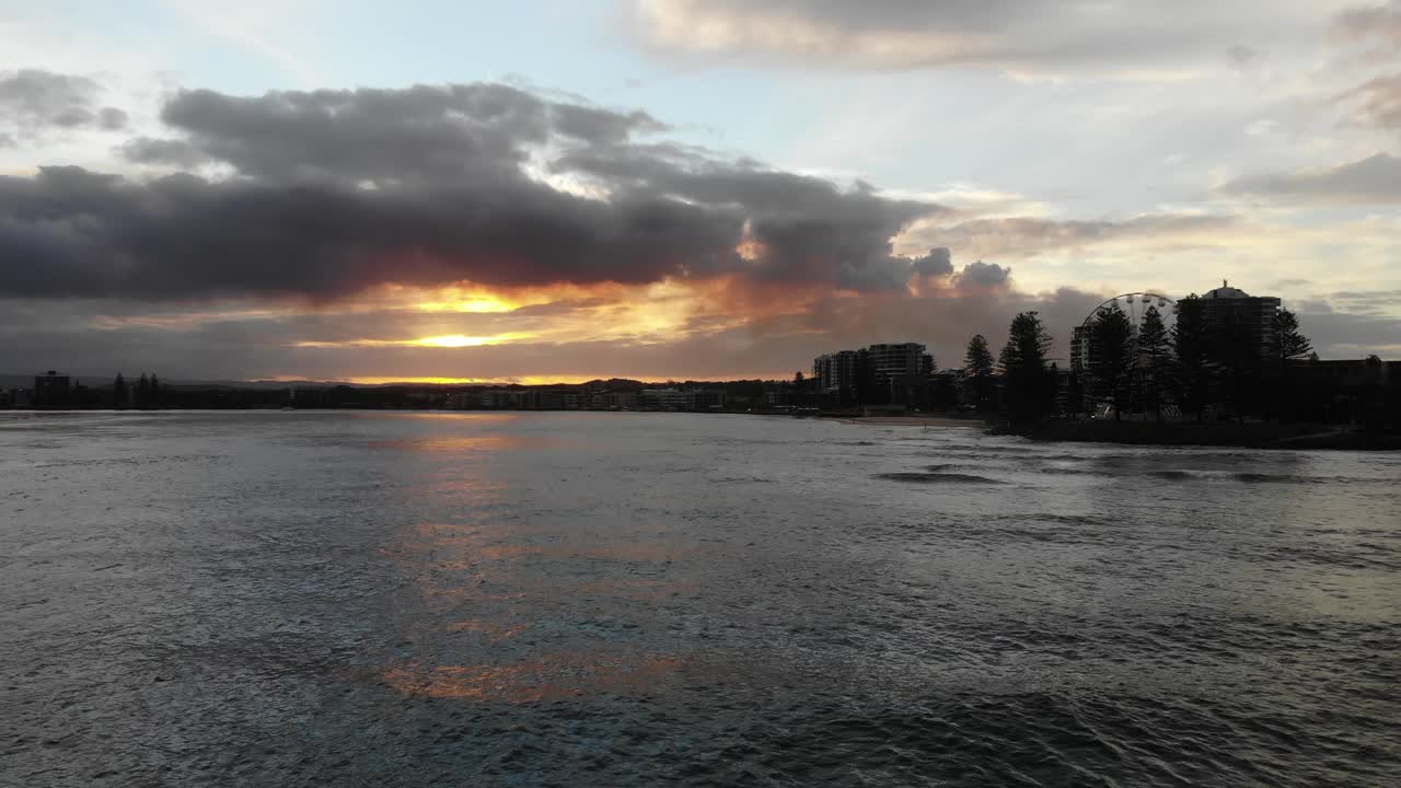 volando lejos de una hermosa puesta de sol ardiente que da luz a un pequeño pueblo de playa que alberga una pequeña rueda de la fortuna escondida detrás de los árboles donde se encuentra una bonita playa a sus pies