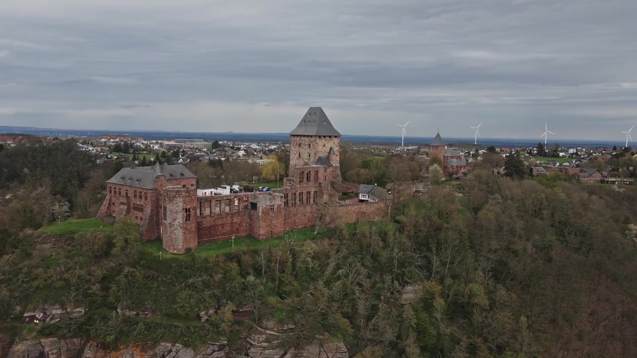 Rotující letecký záběr hradu Nideggen sedícího na okraji útesu vystupujícího ze zeleného lesa. Náladová scéna za zataženého jarního dne v Německu. V pozadí větrné turbíny obnovitelných zdrojů energie