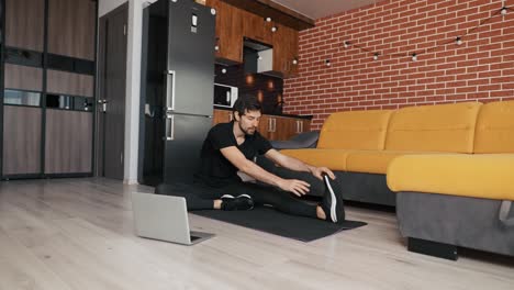 tipo viendo entrenamiento en línea de flexibilidad en una computadora portátil sentado en una alfombra de yoga