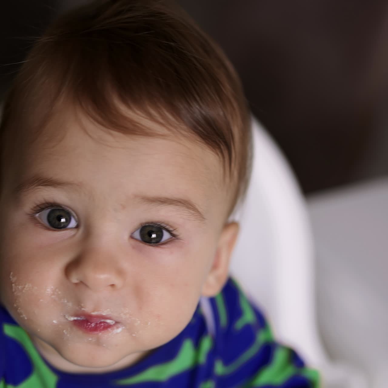 Lovely Caucasian toddler boy holding food in his mouth. Baby with smudged face looking up carefully chewing food. Close up