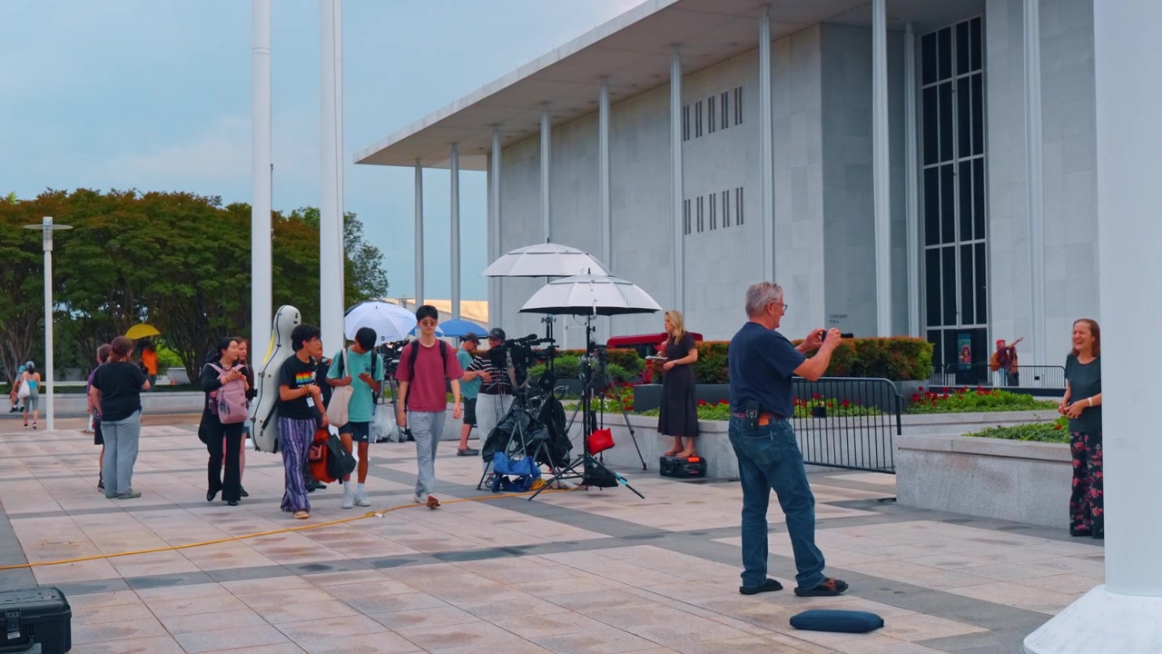 Washington, AS, 13 Juni 2026: Kru televisi sedang syuting di luar Kennedy Center bersama pengunjung. Tripod kamera dan payung pencahayaan di pelataran batu saat musisi muda lewat membawa kotak instrumen
