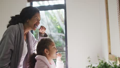 Happy-mixed-race-mother-and-daughter-brushing-teeth-in-bathroom