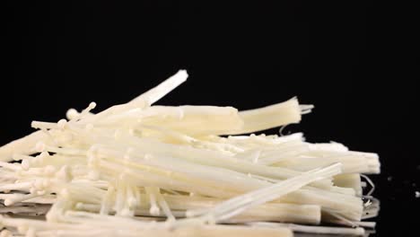 white enoki mushrooms fall and scatter onto a glossy black surface, captured in high-speed close-up with dramatic studio lighting and minimal background