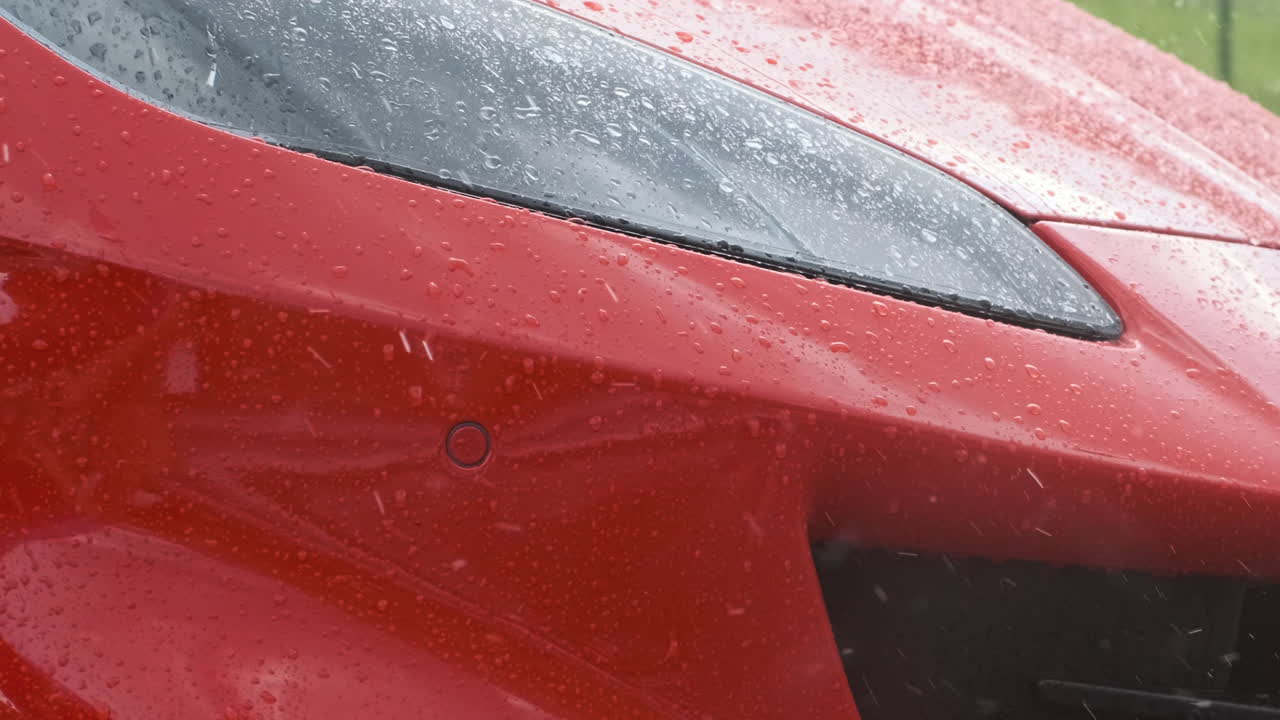 Close up of luxury red sports car slowly reversing in rain. Raindrops cover the headlight and body, cinematic wet weather automotive detail