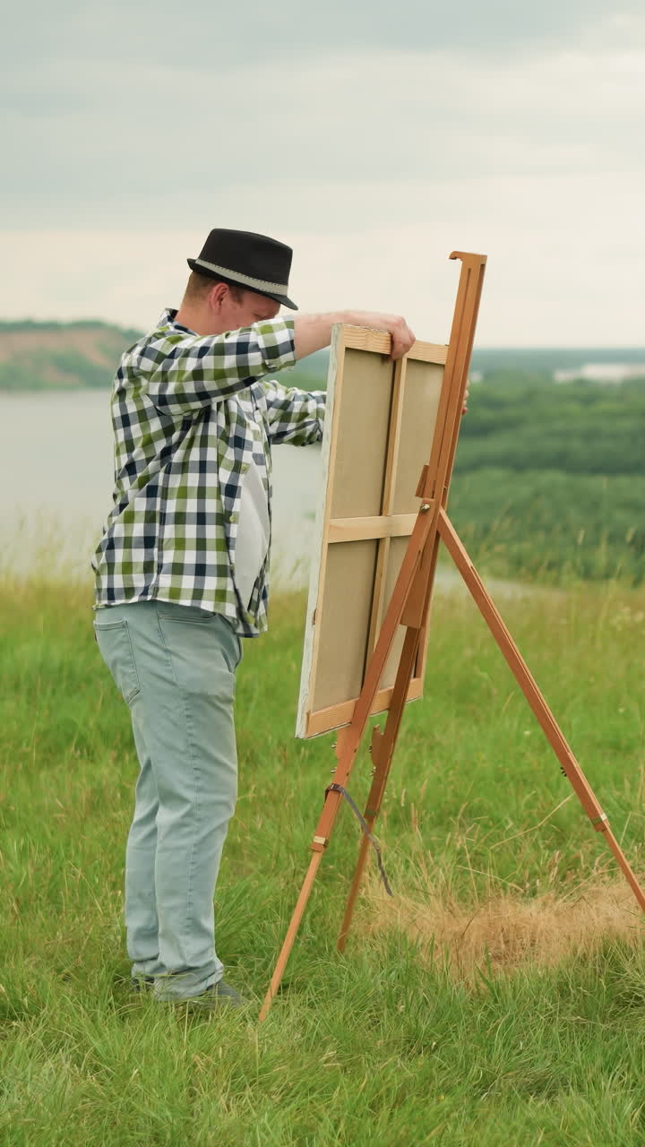 검은 모자와 칠색 셔츠를 입은 예술가가 잔디에 있는 나무 삼각대 위에 그림을 그리는 보드를 세우고 있다.