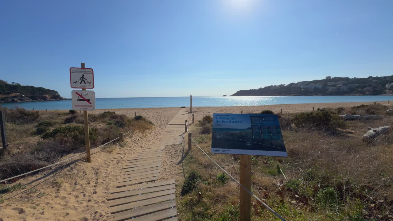 Wooden walkway stretches to a serene beach in girona, lined with signage, under a clear blue sky