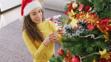 Video-Einer-Glücklichen-Gemischtrassigen-Frau-Mit-Weihnachtsmütze,-Die-Lächelt-Und-Zu-Hause-Den-Weihnachtsbaum-Schmückt