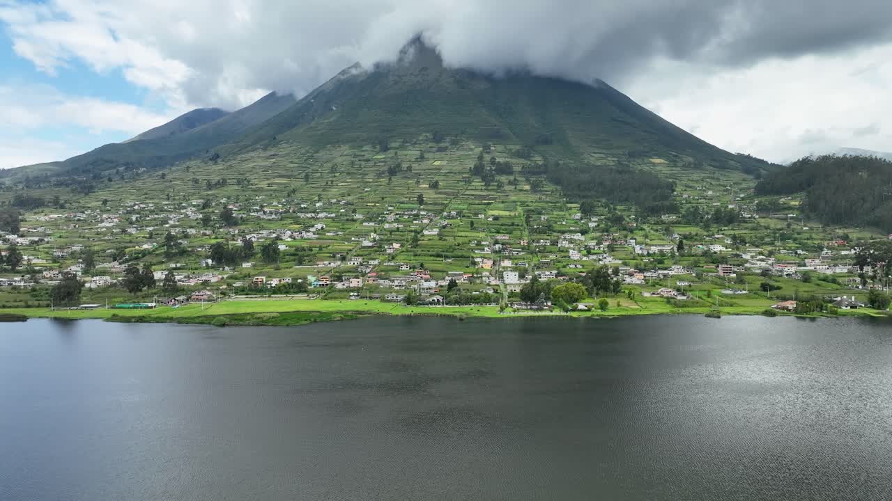Profile view of a town situated at bottom of mountain obscured with clouds and a lagoon in foreground. Drone view.