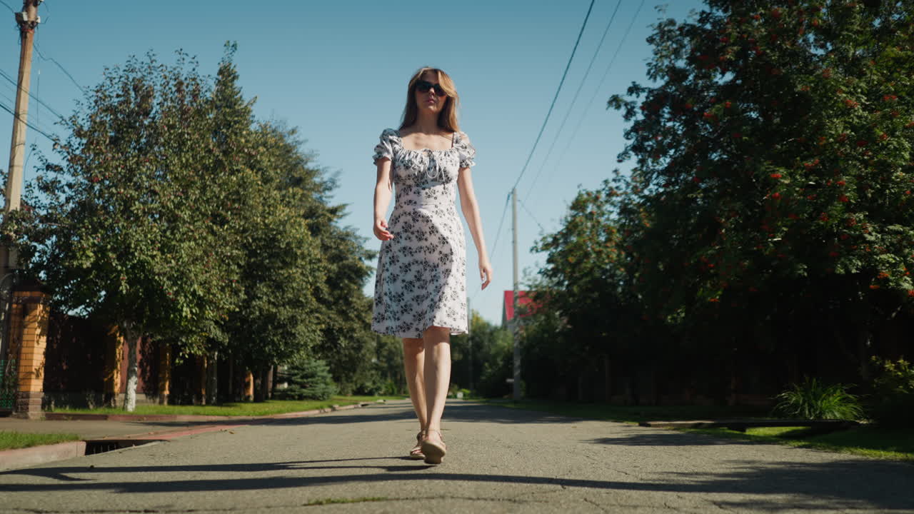 Confident business woman wearing sunglasses walking solo under bright sunlight on quiet street, showcasing floral dress with graceful steps, surrounded by lush trees and suburban tranquility