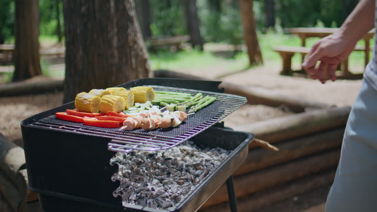 Primo piano di mani maschili che grigliano spiedini di verdure durante un barbecue nella foresta. Persona sconosciuta