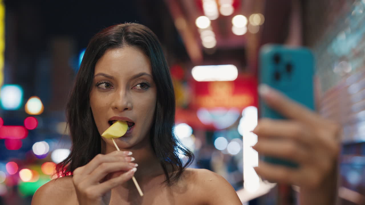 Donna che mangia un mango per strada di notte mentre si fa un selfie.