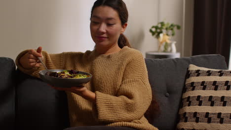 woman at home sitting on sofa eating healthy vegetarian or vegan meal of salad leaves with seeds and dressing with glass of wine watching show on laptop