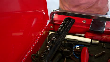 mechanic white caucasian man sets his cell phone android down on red toolbox, grabs socket wrench to work on car, as camera pans down to reveal toolbox with tools and equipment