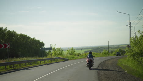 vista trasera de las hermanas montando una bicicleta eléctrica lentamente mientras navegan por una curva en la carretera, una vista borrosa de los coches que vienen de la dirección opuesta, con árboles y un paisaje panorámico en el fondo