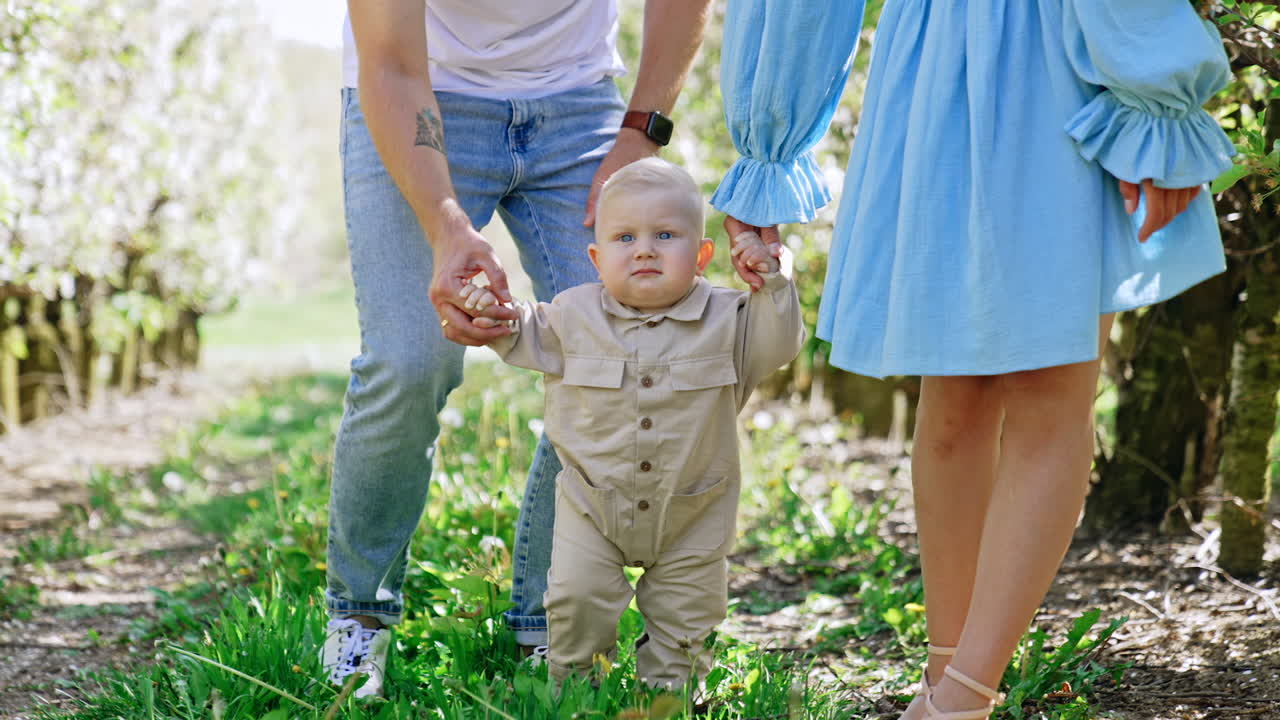 Young parents of a cute toddler learning to walk outdoors. Beautiful Caucasian family portrait in the spring garden.