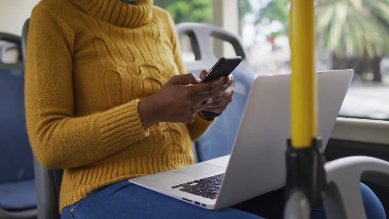 mujer de negocios afroamericana con máscara facial usando un teléfono inteligente y sentada en el autobús