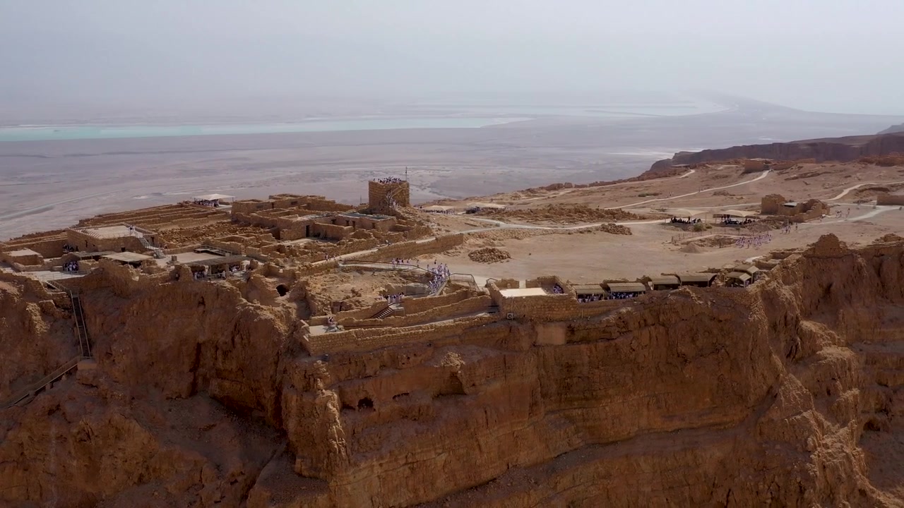 Parlak gündüz gökyüzü altında, uzaktaki Ölü Deniz kıyı şeridinin göründüğü Masada platosundaki tarihi saray yapıları boyunca kaydırılan yüksek açılı drone çekimi.