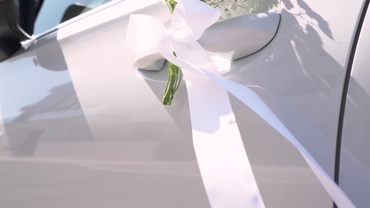 En bil er pyntet med hvite blomster og bånd i et bryllup. Sollyset skinner på bilvinduene mens folk samles rundt arrangementet