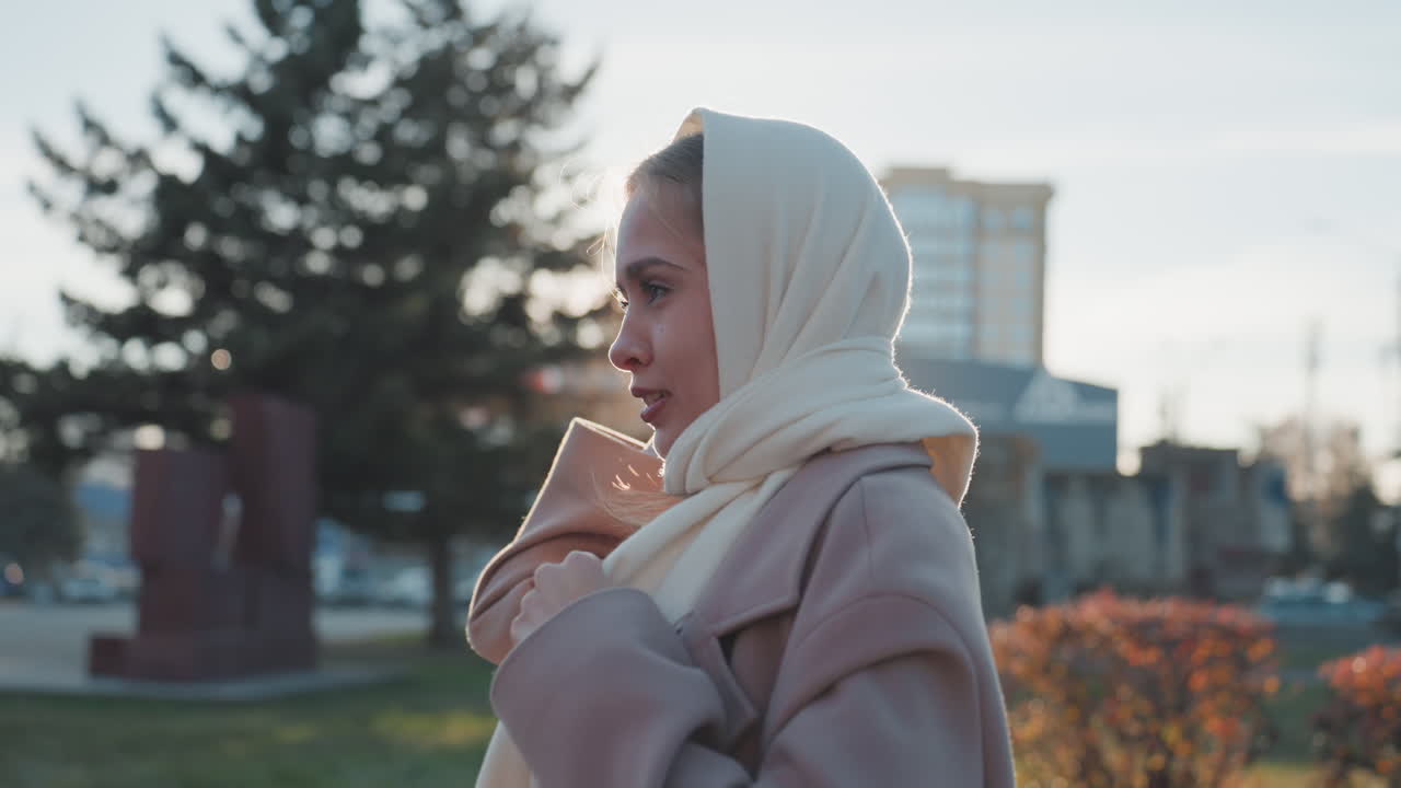 Kremsi başörtülü, yün paltoya sarınmış, altın saatlerde güneşli şehir parkında yürüyen siyah bir kadın, yumuşak arka ışık, düşünceli bir ruh hali, hafif esinti, sonbahar yaprakları, arka planda kamu binaları.