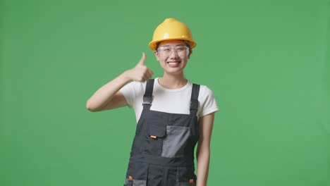 asian woman worker wearing goggles and safety helmet smiling and showing thumbs up gesture while standing in the green screen background studio