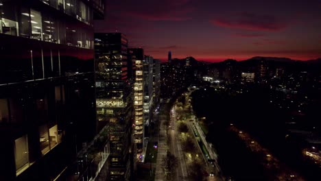 Sobrevuelo-De-Los-Modernos-Edificios-De-Nueva-Las-Condes-En-Santiago-De-Chile,-El-Centro-De-Negocios-De-La-Ciudad,-Con-La-Torre-Costanera-Al-Fondo-Por-La-Noche-Con-Una-Puesta-De-Sol-Naranja