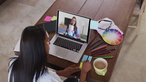 mujer caucásica usando una computadora portátil en una videollamada con una colega, tomando notas